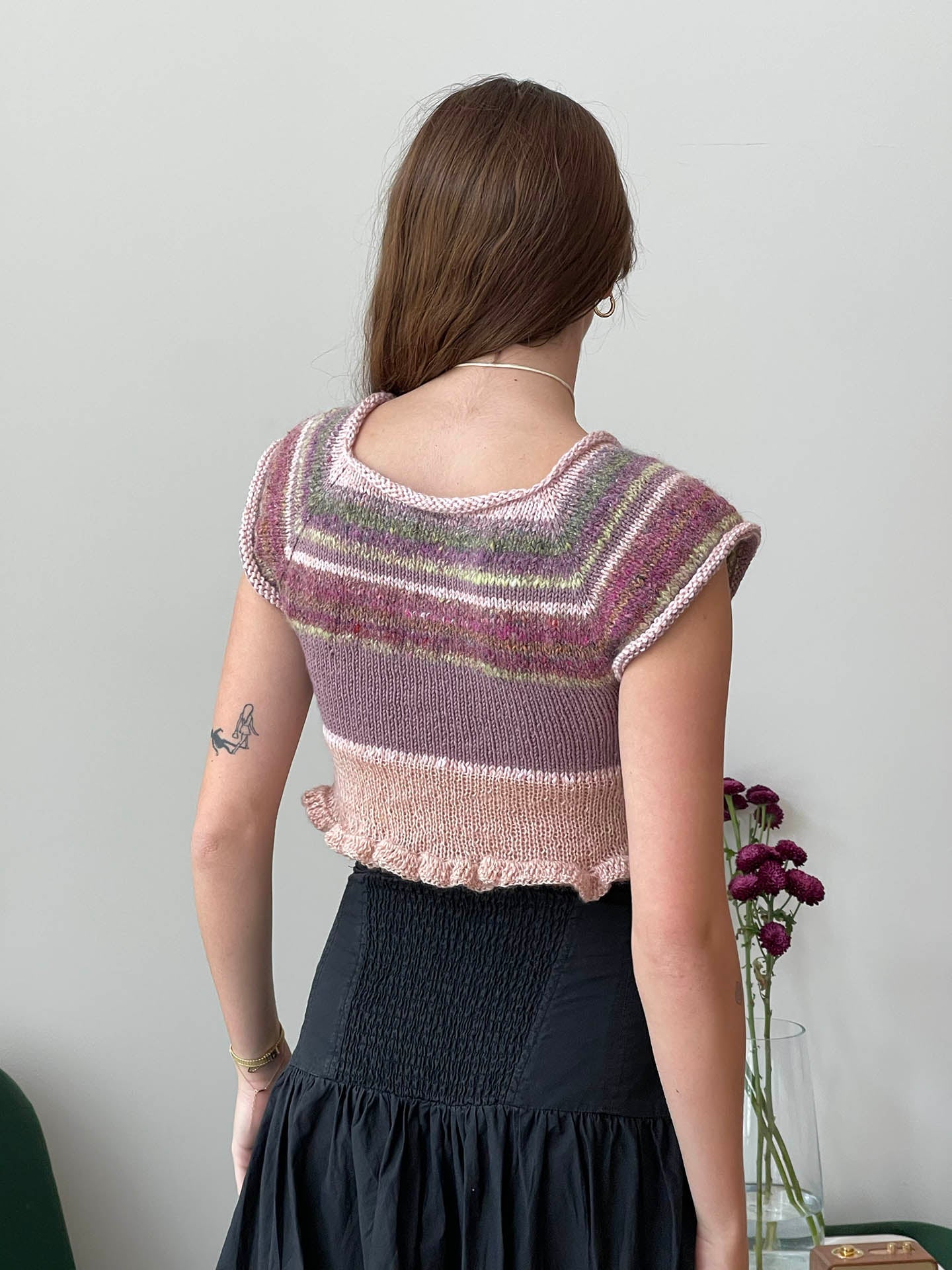 Model turned around showing the back of a cropped knit shirt with hues of pink and purple with a ruffled bottom edge